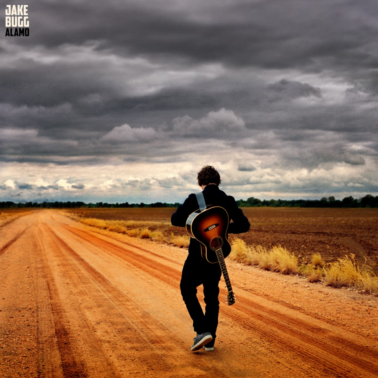 Album cover artwork for Alamo by Jake Bugg, showing the singer walking down a dirt road carrying an acoustic guitar under a stormy sky.